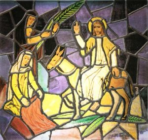 Holy-Week A stain glass picture representing the entering of Christ in Jerusalem riding a donkey while someone waves plam branches and other person lays a cloak on the ground.